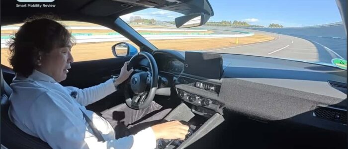 El interior de un coche, específicamente un Honda Prelude 2026, con una persona conduciendo en una pista de carreras. El conductor sostiene el volante con la mano izquierda y parece tener el control del vehículo. El salpicadero y la consola central del coche son visibles, con una pantalla digital y varios controles. La pista de carreras se ve a través del parabrisas, con un cielo despejado y una curva en la pista por delante.