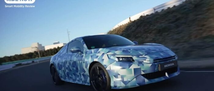Un Honda Prelude 2026 con un envoltorio de camuflaje conduciendo por una carretera. El coche parece ser un prototipo o un vehículo de prueba, como indica el patrón de camuflaje, que se utiliza comúnmente para ocultar los detalles de diseño de nuevos modelos durante las pruebas. El fondo incluye una carretera con barandillas y algunos edificios a lo lejos. La imagen está etiquetada con 'RIDE NOW' y 'Smart Mobility Review' en la esquina superior izquierda, lo que sugiere que es parte de una revisión o contenido promocional relacionado con la movilidad inteligente o la tecnología automotriz.