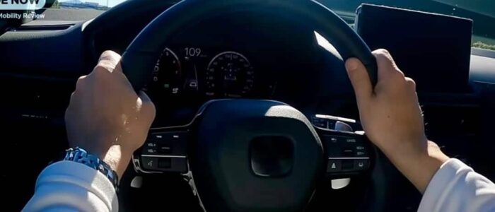 "Una persona conduciendo un Honda Prelude 2026, con ambas manos en el volante. El tablero es visible y muestra una velocidad de 109 km/h. El volante tiene varios controles, incluyendo botones de volumen y control de crucero. También hay una pantalla digital montada en el tablero a la derecha del volante. La imagen parece estar tomada desde la perspectiva del conductor, proporcionando una vista clara de la carretera y los controles interiores del coche.