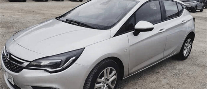 La imagen muestra un Opel Astra plateado estacionado en un aparcamiento al aire libre. El coche está posicionado en un ángulo, mostrando la parte delantera y el lado derecho. El fondo incluye varios otros coches estacionados y algunos edificios con árboles, lo que indica un entorno urbano. Esta imagen es ficticia y se utiliza para un estudio que demuestra las habilidades de marketing de Miana.