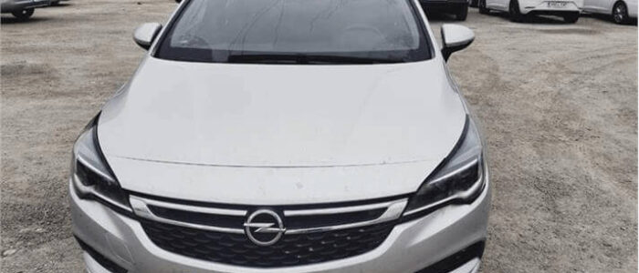 La imagen muestra un Opel Astra plateado estacionado en un aparcamiento. El coche está rodeado de otros vehículos y en el fondo se pueden ver edificios y árboles. La matrícula es parcialmente visible, con la letra "E" indicando que el coche está registrado en España. Esta imagen es relevante ya que proporciona una vista clara del estado y apariencia del coche, lo cual es útil para alguien interesado en comprar un Opel Astra de segunda mano en Zaragoza.