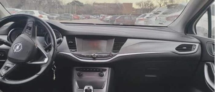 La imagen muestra el interior de un Opel Astra de segunda mano en Zaragoza. Se pueden ver los asientos delanteros, el salpicadero y el volante. El coche está estacionado en un aparcamiento con otros vehículos visibles a través del parabrisas. El salpicadero cuenta con una pantalla central, rejillas de ventilación y varios controles. El volante tiene el logotipo de Opel en el centro y hay una palanca de cambios en la consola central. Los asientos tienen un tejido con patrón. Esta imagen es ficticia y se utiliza para un estudio que demuestra las habilidades de marketing de Miana.