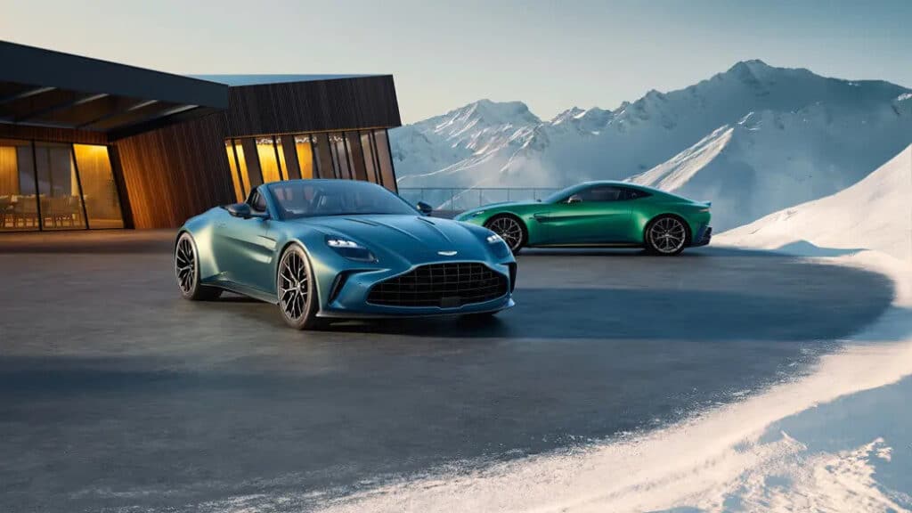 Imagen de un Aston Martin Vantage Roadster 2025 en un entorno pintoresco. El coche descapotable es de color azul metálico, con líneas elegantes y un diseño aerodinámico. El techo de lona está plegado, mostrando el interior lujoso del vehículo. Las llantas de aleación y los faros LED destacan en la imagen, realzando la apariencia deportiva y sofisticada del coche.