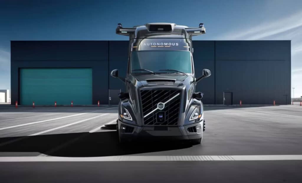 Un camión Volvo autónomo de color plateado se encuentra estacionado en la calle. No hay conductor visible en la cabina y el camión está equipado con sensores y cámaras en el techo, indicando su capacidad de conducción autónoma. Su diseño robusto y moderno destaca.