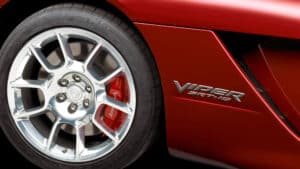 Primer plano de la rueda delantera de un coche deportivo rojo, con llanta de aleación de múltiples radios y freno de disco con pinza roja. En la carrocería se distingue el emblema 'Viper SRT-10', identificando al vehículo como un Dodge Viper de alto rendimiento.