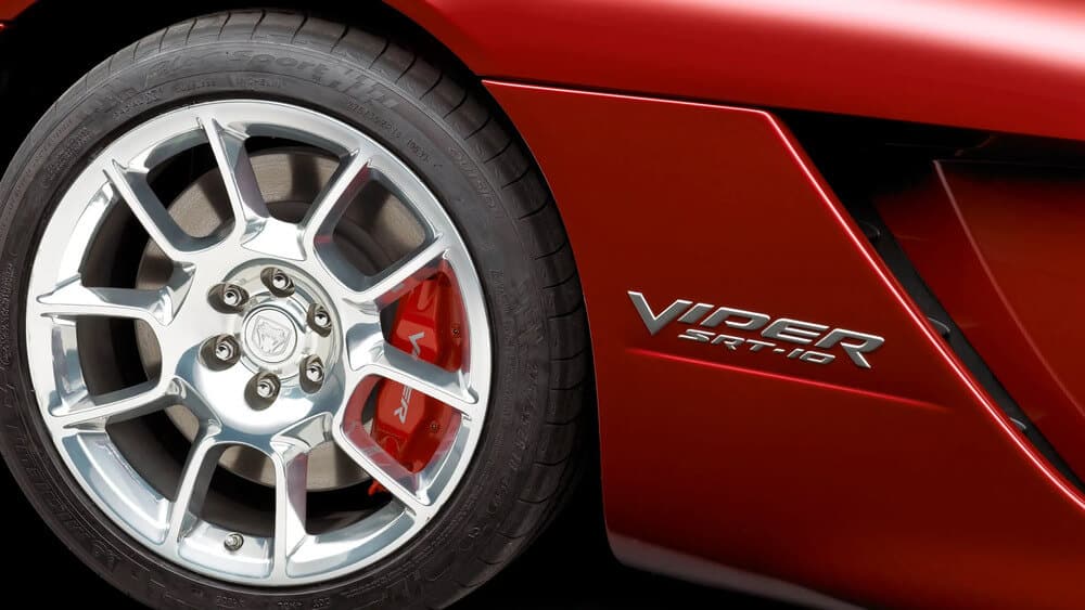 Primer plano de la rueda delantera de un coche deportivo rojo, con llanta de aleación de múltiples radios y freno de disco con pinza roja. En la carrocería se distingue el emblema 'Viper SRT-10', identificando al vehículo como un Dodge Viper de alto rendimiento.